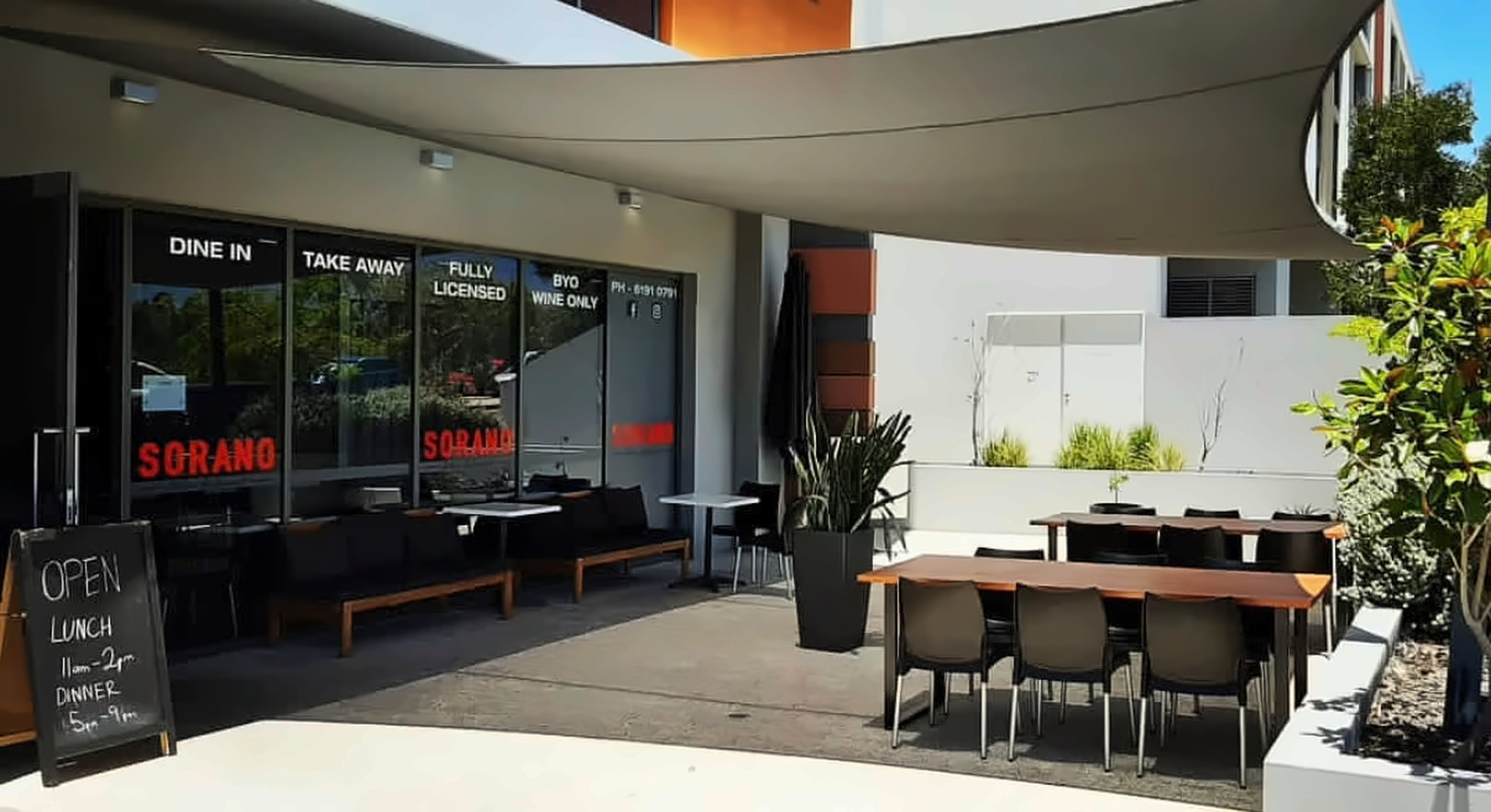 Sorano's sunny courtyard under a shade sail, with timber tables and windows lettered SORANO, dine in, take away and fully licensed