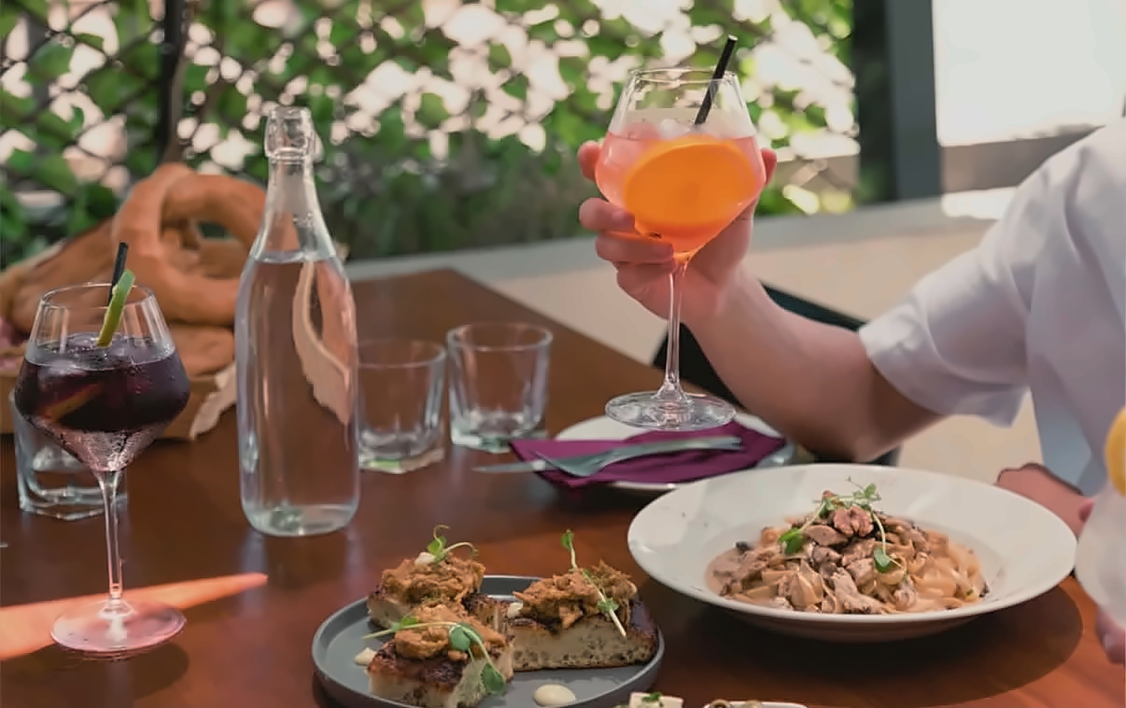 A shared table at Sorano with a spritz raised, bruschetta, a bowl of creamy pasta, sangria and a carafe of water